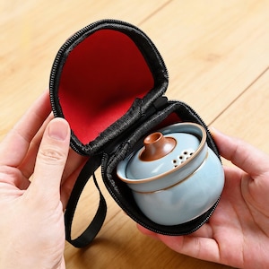 May include: A small, light blue ceramic tea set with a brown lid is nestled inside a black zippered carrying case with a red interior. The tea set is round and has a series of small holes near the top.
