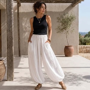 Könnte beinhalten: Eine Frau trägt ein schwarzes Tanktop und eine weiße Leinenhose. Die Hose hat einen lockeren Schnitt mit gerafften Knöcheln. Sie trägt braune Sandalen und eine Halskette. Im Hintergrund ein Baum im Topf und eine Holzpergola.
