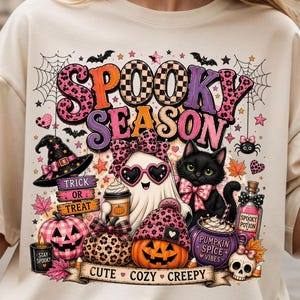 May include: A cream-colored t-shirt featuring a graphic print with the text "SPOOKY SEASON" and "CUTE COZY CREEPY". The design includes a ghost with heart-shaped glasses, a black cat, a witch hat, pumpkins, a potion bottle, and a skull.