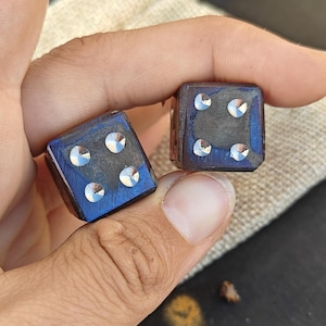 Puede incluir: Dos dados cuadrados con un acabado metálico iridiscente en azul oscuro y violeta. Cada cara muestra cuatro puntos plateados circulares y reflectantes dispuestos en un patrón cuadrado. Los dados tienen bordes definidos y superficie lisa.