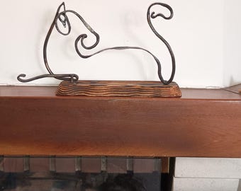 Pode incluir: Uma escultura de arame de metal escuro que representa o contorno abstrato de um gato em posição sentada. A escultura está montada sobre uma base de madeira retangular com veios horizontais, acabamento escuro queimado e textura visível.