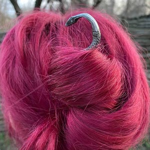 Könnte beinhalten: Eine dunkelgraue, handgeschmiedete Haarnadel aus Metall mit einem gebogenen, hakenförmigen Kopf. Die Haarnadel steckt in einem Dutt aus leuchtend magentafarbenem Haar. Die Metalloberfläche weist sichtbare Hammerschläge und ein rustikales Finish auf.