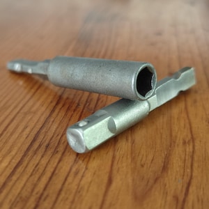 Może przedstawiać: Dwie srebrne metalowe przedłużki do wierteł. Jedna przedłużka posiada na końcu sześciokątny otwór nasadowy, a druga ma kwadratową końcówkę napędową. Oba narzędzia mają sześciokątny chwyt przeznaczony do montażu w wiertarce elektrycznej.
