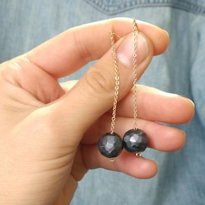 May include: A pair of gold-colored chain earrings, each with a dark blue, faceted sphere pendant. The earrings are displayed against a light blue denim background.