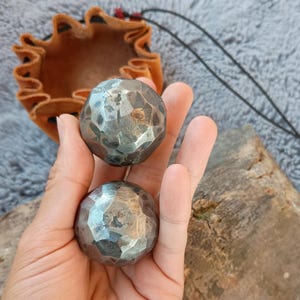 May include: Two textured, metallic spheres with a hammered appearance are held in a hand. A small, brown leather pouch with a drawstring is in the background. The spheres have a faceted surface, reflecting light. The pouch is open, revealing its interior.