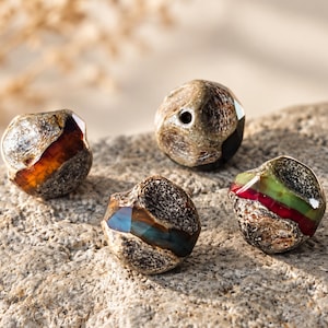 May include: Four spherical beads with a rough, textured, greyish-brown exterior and smooth, polished, multi-coloured bands. The beads feature translucent sections in amber, deep blue, and green-red tones, each with a central hole for threading.