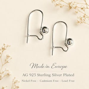 Op de afbeelding: Een paar zilverkleurige oorbellen met een gebogen haakontwerp en een enkele gepolijste bolvormige kraal aan de zijkant. De tekst luidt: Made in Europe, AG 925 Sterling Silver Plated, Nickel Free, Cadmium Free, Lead Free.