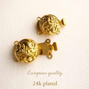 Puede incluir: Dos cierres dorados con un diseño floral ornamentado. Los cierres redondos tienen una superficie texturizada y una pequeña pieza rectangular con agujeros para la fijación. El texto "European quality" y "24k plated" está debajo.