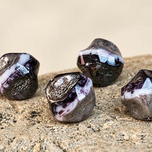 May include: Four faceted glass beads featuring a marbled pattern of deep purple, black, and white. Each bead has a polished, multi-faceted surface with metallic silver-toned accents integrated into the dark, swirling glass composition.