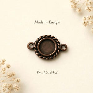 Puede incluir: Un conector de joyería redondo de metal color cobre con un borde de cuerda trenzada y un engaste circular central. El componente incluye dos pequeñas anillas en los lados opuestos y muestra el texto Made in Europe y Double-sided.