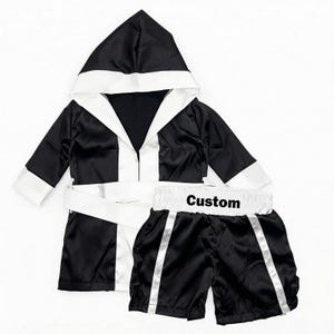 Könnte beinhalten: Schwarzes und weißes Box-Robe- und Shorts-Set. Die Robe hat eine Kapuze und weiße Verzierungen. Die Shorts haben weiße Streifen und das Wort "Custom" auf dem Bund. Das Set ist aus glänzendem Material.