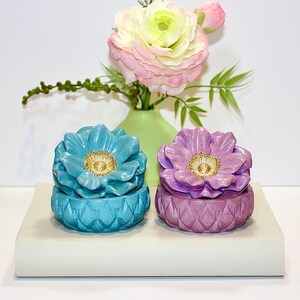 May include: Two ceramic flower-shaped trinket boxes, one turquoise and one lavender, with gold-colored centers. The boxes sit on a white surface, with a vase of flowers in the background. The flowers are pink and yellow.