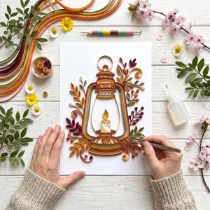 Könnte beinhalten: Ein Papier-Quilling-Kunstwerk auf weißem Papier mit einer zentralen Laterne und einer brennenden Kerze, umgeben von kunstvollen Papierrollen in Orange-, Rot-, Braun- und Gelbtönen, angeordnet mit dekorativen Blättern und floralen Elementen.