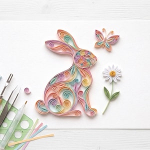 May include: A colorful paper quilling art piece featuring a rabbit, butterfly, and daisy. The rabbit is made of pastel-colored paper strips, with a butterfly and daisy nearby. Crafting tools and paper strips are visible.