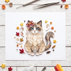 May include: A paper quilling art piece featuring a fluffy cat with blue eyes and a purple and orange bow. The cat is surrounded by paper quilled elements including yellow stars, red and brown leaves, and various swirls in shades of cream and tan.