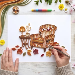 May include: A paper quilling art piece on white paper featuring an armchair, a side table with a lamp and a steaming mug, and scattered autumn leaves. The design uses coiled paper strips in shades of orange, brown, and yellow.