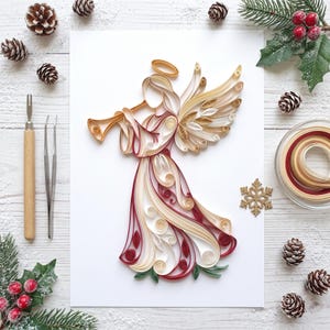 May include: A paper quilling art piece depicting an angel playing a trumpet, crafted from coiled strips of cream, burgundy, and gold paper. The angel features a halo, wings, and a flowing gown, displayed on a white rectangular cardstock base.