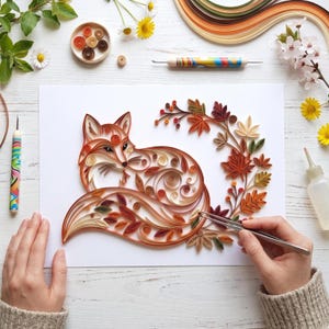 Könnte beinhalten: Ein Papier-Quilling-Kunstwerk auf weißem Papier, das einen Fuchs aus gerollten Papierstreifen in Orange-, Braun- und Cremetönen zeigt, umgeben von einem dekorativen Kranz aus Herbstblättern und Beeren, zusammen mit Bastelwerkzeugen und Blumen.