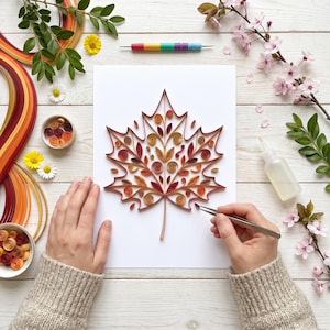 Könnte beinhalten: Ein Papier-Quilling-Kunstprojekt mit einem Ahornblatt-Design aus gerollten Papierstreifen in Rot-, Orange- und Brauntönen auf einem weißen Blatt Papier, umgeben von Quilling-Werkzeugen, Papierstreifen und floralen Elementen.
