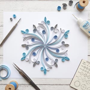 May include: A paper quilling snowflake design made of curled strips in shades of light blue, dark blue, white, and gray. The snowflake is surrounded by quilling tools, a bottle of quilling glue, a circle sizing template, and spools of paper.