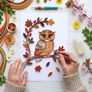 Könnte beinhalten: Ein Papier-Quilling-Kunstprojekt auf weißem Papier mit einer braunen Eule, die auf einem Ast mit Herbstblättern sitzt. Die Szene umfasst bunte Papierstreifen, ein Quilling-Werkzeug, eine kleine Klebeflasche und lose Papierrollen.