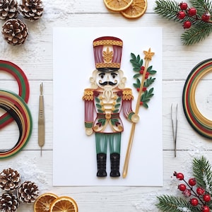 Könnte beinhalten: Ein Papier-Quilling-Kunstwerk, das einen Nussknacker-Soldaten in Rot, Grün, Gold und Schwarz darstellt. Die Figur trägt einen hohen Hut, eine detaillierte Uniform und hält einen Stab mit einem Stern, umgeben von Tannenzapfen und Orangenscheiben.