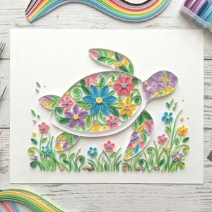 Op de afbeelding: Een kleurrijk papier quilling kunstwerk van een zeeschildpad, versierd met bloemen in roze, blauwe, gele en paarse tinten. De schildpad is omgeven door groen gras en bloemen, met knutselgereedschap en papieren stroken op de achtergrond.