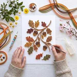 Könnte beinhalten: Ein Papier-Quilling-Kunstprojekt auf weißem Papier mit detaillierten Eichenblättern und Eicheln in Orange-, Braun-, Gelb- und Grüntönen. Die Komposition umfasst gebogene Papierstreifen, ein Quilling-Werkzeug, eine Pinzette und Kleber.