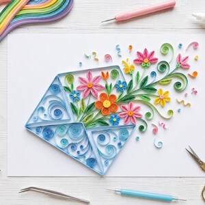 May include: A paper quilling art piece on white paper featuring a blue envelope shape filled with colorful paper flowers in pink, orange, and blue, surrounded by green paper leaves and swirls, with crafting tools like scissors and quilling pens nearby.