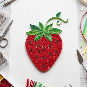 May include: A handmade paper quilling strawberry with green leaves and stem, set against a white background. The red fruit features dark brown seeds, and is surrounded by craft tools and colorful paper strips.