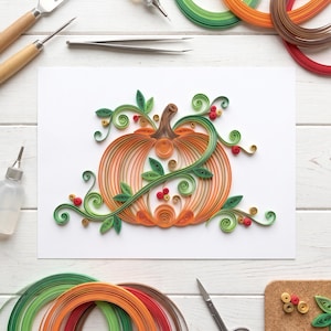 May include: A paper quilling art piece on white paper featuring an orange pumpkin with a brown stem, surrounded by green swirling vines, leaves, and small red and yellow berries. Crafting tools and colorful paper strips are arranged around it.