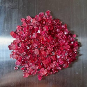 May include: A pile of raw, uncut ruby gemstones. The rubies are a deep, translucent red color, with irregular shapes and facets. The gemstones are scattered on a metallic surface.