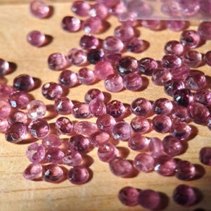 以下が含まれることがあります： 表面に散らばった、小さく丸いファセットカットのピンク色の宝石のコレクション。個々の石は半透明で、複数のカット面が光を反射し、さまざまな色合いのピンク色で輝きを放っています。