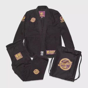 Puede incluir: Conjunto de uniforme de artes marciales negro con logotipos bordados en oro y morado. Incluye una chaqueta, pantalones y una bolsa con cordón. La chaqueta y los pantalones tienen la marca "Shoyoroll".