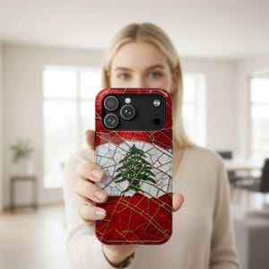May include: A red, white, and green phone case featuring the Lebanese flag design. The case has a mosaic-like pattern with a green cedar tree in the center. The case is held up in front of a person.
