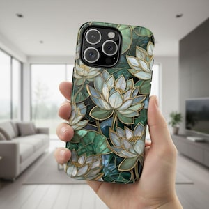 May include: A smartphone case featuring a floral design with white, gold, and green lotus flowers. The case has a dark green background and a glossy finish. The phone case is held in a hand.