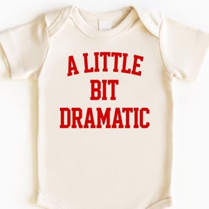 May include: Cream-colored baby onesie with the text "A LITTLE BIT DRAMATIC" in bold red letters. The onesie has short sleeves and snaps at the bottom for easy changes. A cute and humorous baby garment.