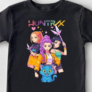 May include: Black t-shirt featuring a colorful graphic of four anime-style characters and a blue cat. The word "HUNTRIX" is displayed above the characters in a rainbow font. The design includes hearts and stars.