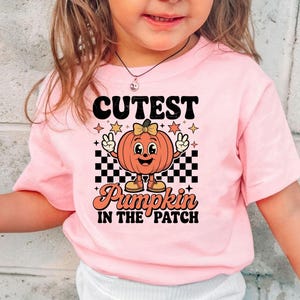 May include: Pink short-sleeved t-shirt featuring a retro-style pumpkin graphic with the text "Cutest Pumpkin in the Patch." The pumpkin illustration includes a bow, shoes, and peace signs.