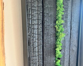May include: A wall art piece featuring vertical planks of charred black wood with a deep, cracked texture. A single, continuous strip of vibrant, bright green preserved moss runs vertically between two of the dark, burnt wooden boards.