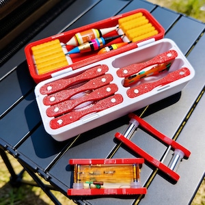 May include: A collection of fishing tackle, including red and white tackle boxes, floats, and other fishing accessories. The tackle boxes are open, revealing the contents. The image is taken outdoors.