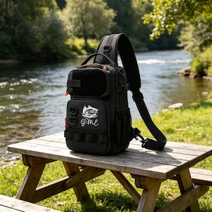 May include: Black sling bag with orange accents, resting on a wooden picnic table. The bag features a front pocket with a hook-and-loop patch, multiple attachment points, and a graphic of a fish jumping out of water with the word "gme".