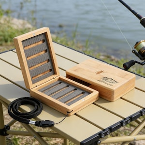 Może przedstawiać: Dwa otwarte drewniane pudełka do łowienia much z szarymi wkładkami z pianki leżą na beżowym stole. Obok otwartych pudełek znajduje się zamknięte pudełko z grafiką ryby. Widoczna jest również czarna smycz i wędka.