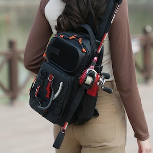 May include: Black fishing backpack with multiple compartments and orange accents. A red and black fishing rod is attached to the side, along with a red fishing reel. The backpack is worn over the shoulder.