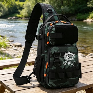 May include: A black and green camouflage sling bag with orange accents. The bag features multiple compartments, a shoulder strap, and a fishing-themed logo with the initials "gMe". The bag is designed for outdoor activities.