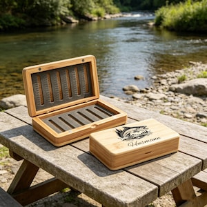 Puede incluir: Una caja rectangular de bambú para pesca con mosca con tapa abatible. La caja abierta muestra un interior de espuma gris oscuro con filas ranuradas para anzuelos. La caja cerrada presenta un grabado láser negro de un pez y el nombre Husmann.