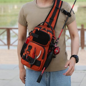 May include: An orange and black fishing tackle shoulder bag with multiple compartments and a rod holder. The bag has a padded shoulder strap and is designed to carry fishing gear. A red and black fishing rod is attached to the bag.
