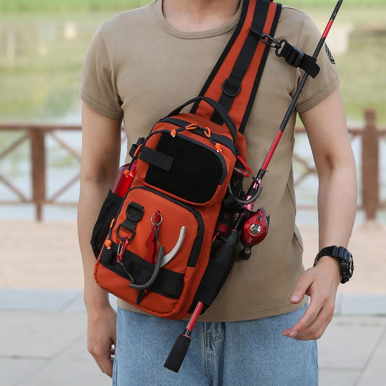 May include: An orange and black fishing tackle shoulder bag with multiple compartments and a rod holder. The bag has a padded shoulder strap and is designed to carry fishing gear. A red and black fishing rod is attached to the bag.