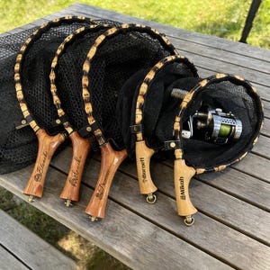 Puede incluir: Cinco redes de pesca con mangos de madera y redes negras se exhiben sobre una mesa de madera. Los mangos están grabados con los nombres: Blanche, Dominic, Barnett y Mikah. Una red contiene un carrete de pesca.