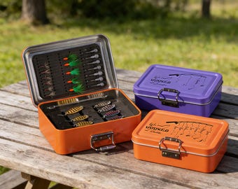 Könnte beinhalten: Eine offene orangefarbene Metall-Angelbox mit Angelködern, neben zwei geschlossenen Boxen, eine lila und eine orangefarbene. Die offene Box hat ein schwarzes Innere mit verschiedenen Angelködern. Die geschlossenen Boxen haben den Text "We're Hooked" und eine Angelruten-Grafik.